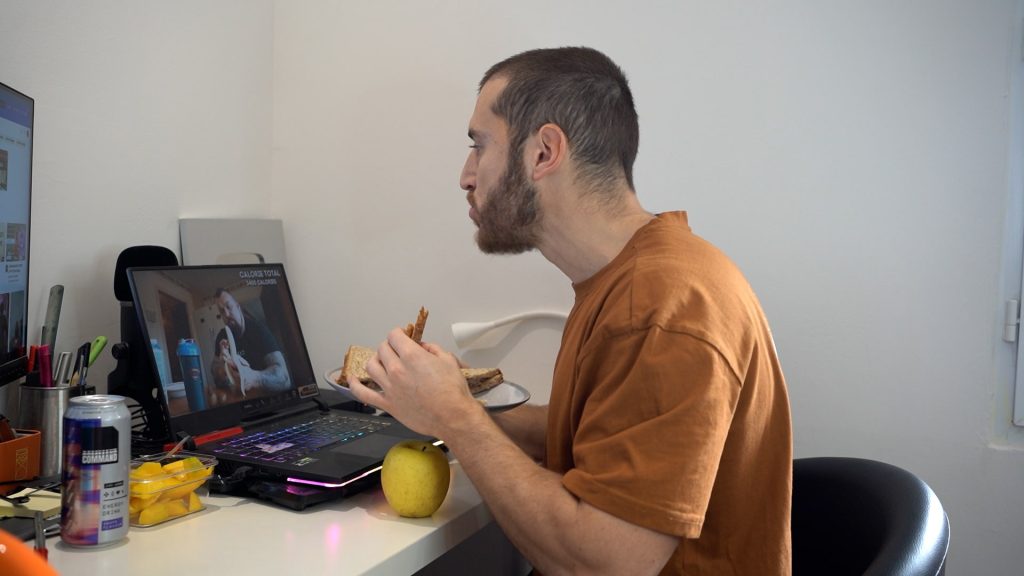 Homme de profil mangeant un sandwich au pâté assis devant son ordinateur avec une pomme à sa gauche