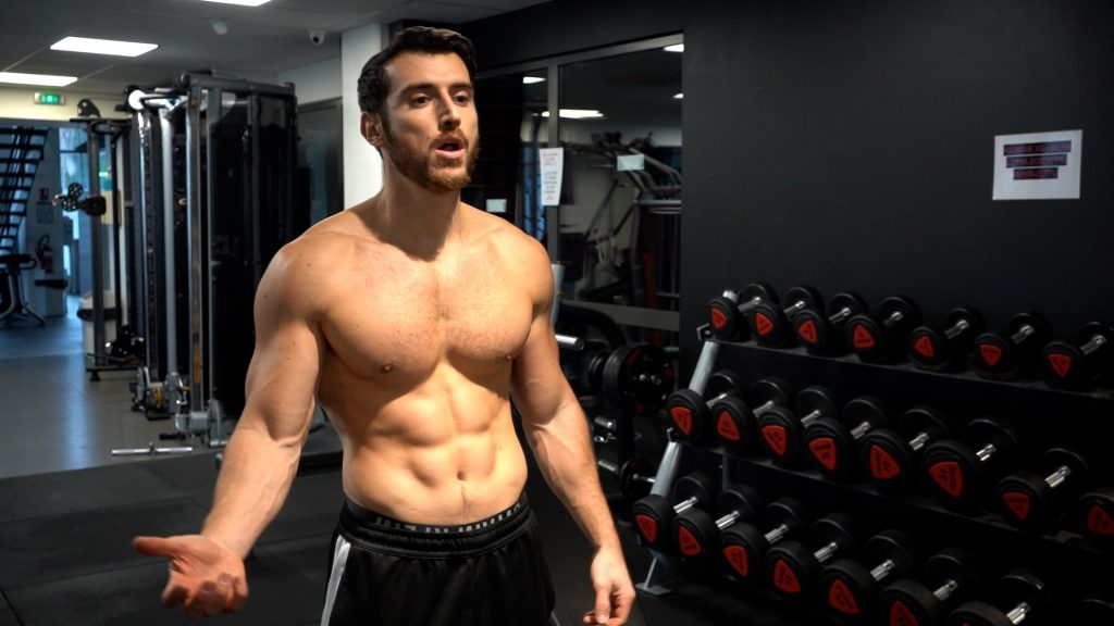 Homme en train de poser torse nu dans un gym avec des haltères et des poulies en fond